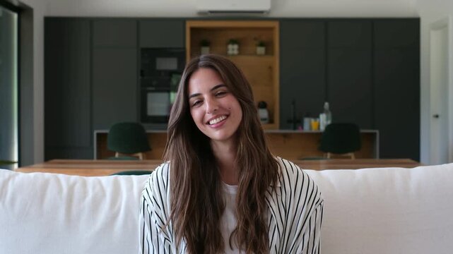 Portrait of a beautiful young woman with long brown hair smiling genuinely at the camera while sitting comfortably on a white sofa in her modern and spacious living room at home