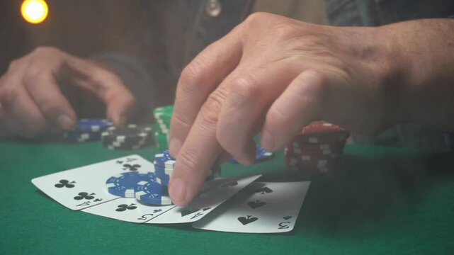 Hands of a male poker player placing blue chips on a table with playing cards and various colored poker chips in a dimly lit setting
