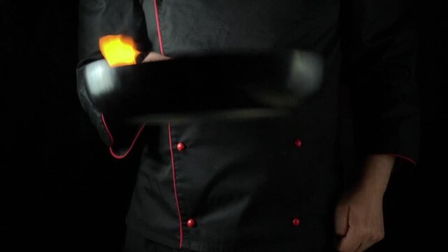 Chef in black uniform skillfully tosses vegetables in a flaming pan, showcasing culinary technique in a dark kitchen setting