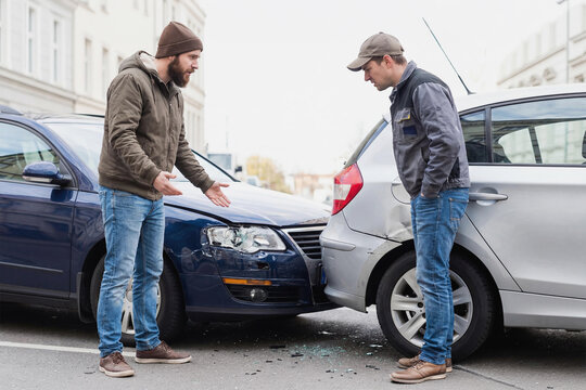 Two men argue after a car accident on a city street, looking at the damage to their vehicles. Image for insurance, road safety, and urban traffic dispute resolution concept.