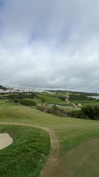 Scenic view of a golf course with rolling hills, sandy bunkers, and coastal landscape under a cloudy sky