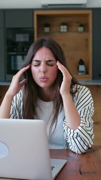Young stressed freelance woman suffering from a painful headache and migraine while working on her laptop from home, feeling exhausted, overworked, and tired from her demanding remote job