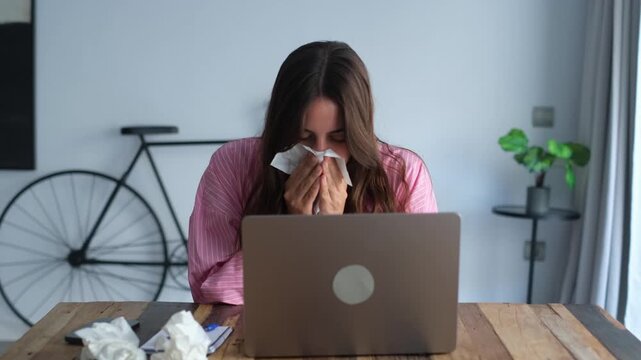 Unhealthy young woman suffering from cold, flu, or allergies, sneezing into a tissue and feeling unwell while trying to work remotely on her laptop computer at her desk in a modern apartment