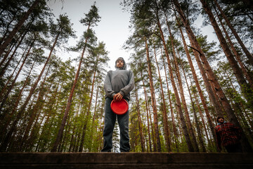 Male disc golfer stands on a wooden platform holding a red frisbee in a dense forest with tall pine trees and a cloudy sky in the background © Emvats