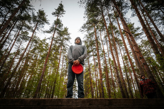 Male disc golfer stands on a wooden platform holding a red frisbee in a dense forest with tall pine trees and a cloudy sky in the background