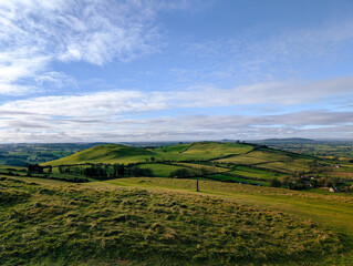 Obraz premium Rolling green hills and fields stretch across the Irish countryside under a bright blue sky with soft clouds