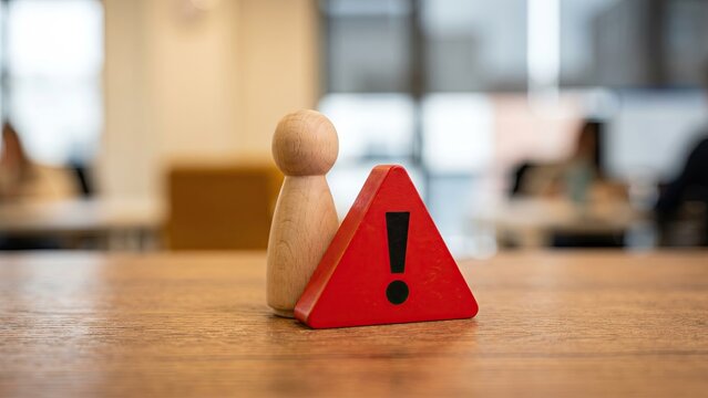 A wooden peg figure standing behind a small red warning triangle on a desk, symbolizing crisis management, risk alert and business continuity.