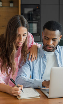 Diverse couple collaborating at home office, woman leaning over partner&rsquo;s shoulder pointing at laptop while he takes notes in a spiral notebook, focused on planning and budgeting