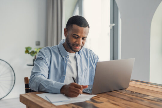 Young black man working on a laptop, writing notes on paper, sitting at a wooden table in a brightly lit home office, focused on remote work and productivity