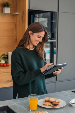 Young woman smiling while browsing a digital tablet in a modern kitchen, preparing a healthy breakfast of toast and orange juice and enjoying a relaxed morning routine
