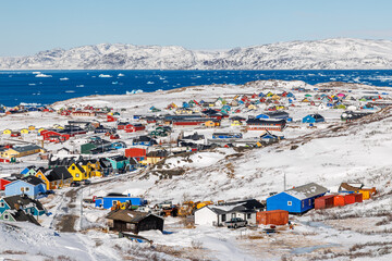 Colorful arctic residential neighborhood with bright wooden Inuit houses scattered across snowy hills above the coast of winter fjord full of icebergs, Narsaq, Kujalleq municipality, Greenland © vadim.nefedov