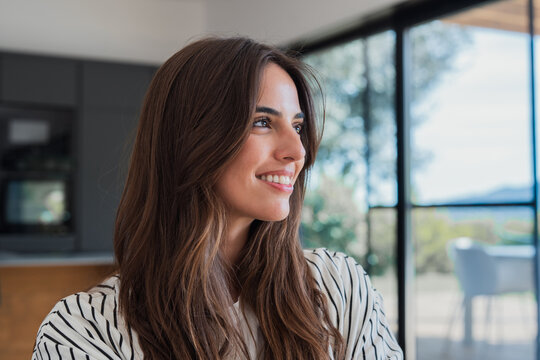 Young smiling woman with long brown hair naturally lit, finding joy and optimism while looking ahead, embodying happiness and success in a modern indoor living space