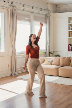 Happy young woman joyfully dancing and singing with raised arm, feeling freedom and positive emotions while enjoying music in her bright living room at home