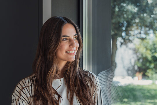 Young woman smiling while looking out a large window, contemplating the future with a hopeful and optimistic expression, enjoying natural light indoors