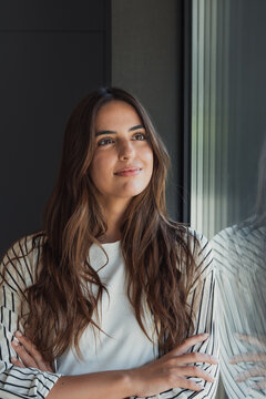 Young woman with long brown hair stands by a window with arms crossed, smiling softly and looking up, daydreaming about future possibilities with calm confidence and peace of mind