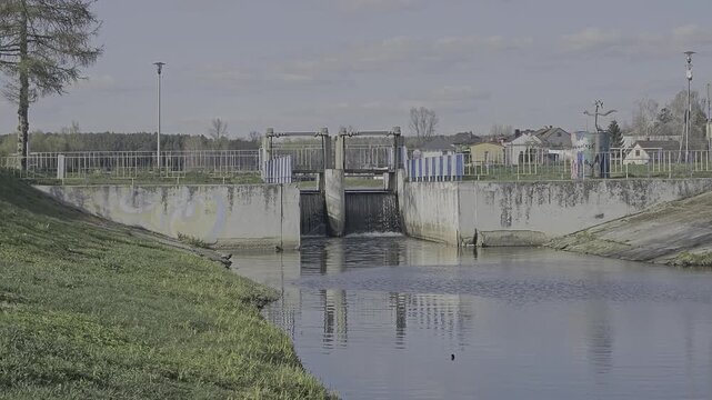 Small concrete water dam with sluice gate on a river in a suburban area, water flowing through the weir, irrigation system infrastructure in a village with houses in the background.