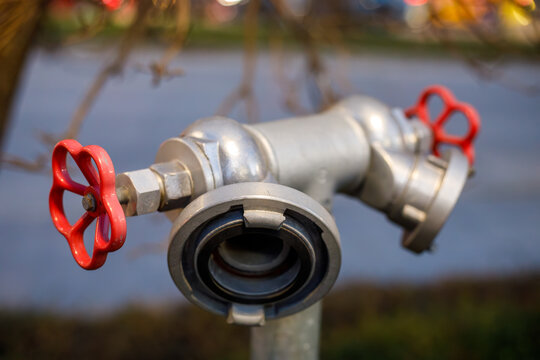 a silver metal fire hydrant standpipe with two red wheel valves and a hose coupling stands on an outdoor post with a blurred background of water and trees