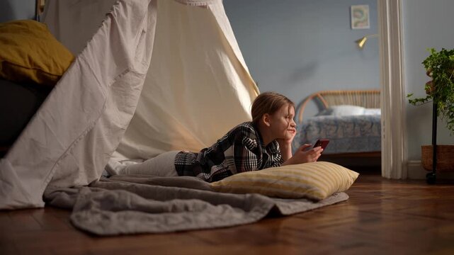 Young caucasian girl smiling in blanket fort replying to phone, relaxed on stomach with yellow pillow and soft blanket, warm evening light, content expression, .