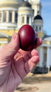 Hand holding a red Easter egg in front of a church, symbolizing preparation for a religious holiday and traditional blessing of Easter eggs. Concept of cultural rituals