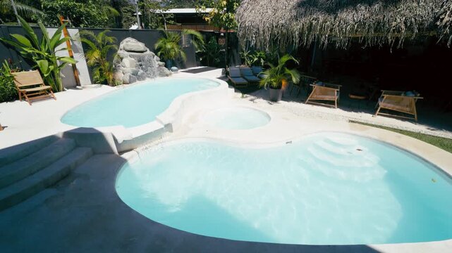 Beautifully designed swimming pool and jacuzzi at a tropical resort hotel. Lush green plants, lounge chairs, and a tiki hut create a serene oasis