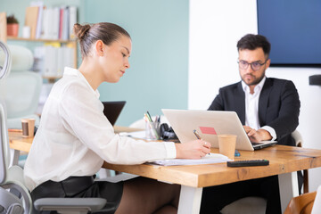 Obraz premium Man and woman working in office, manager writes on paper or fills out contract forms, using laptop in work. Regional representative office company, carsharing, hiring staff.