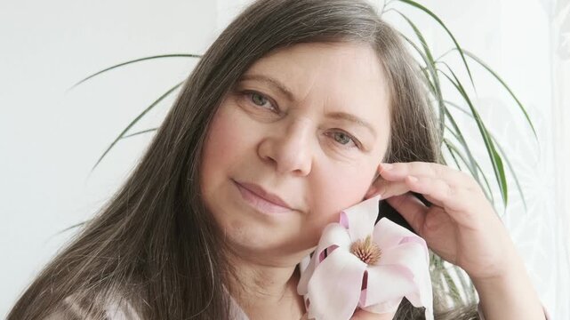 aging woman with magnolia flower looks herself in mirror, noticing wrinkles, changes in facial contour and sagging eyelids, cosmetic anti-aging procedures, natural process growing older