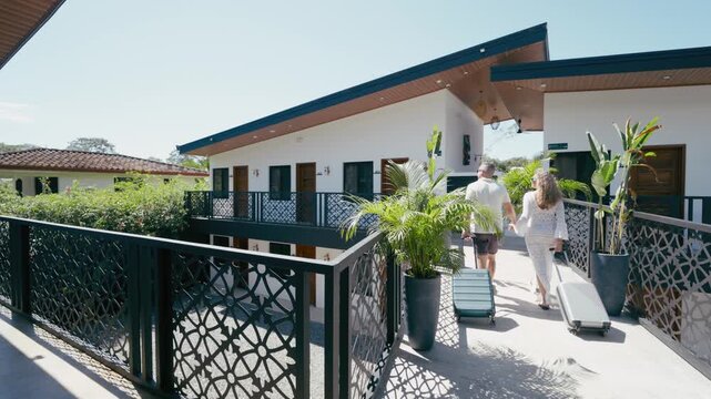 Rear view of a man and woman with rolling suitcases walking along the sunny balcony of a modern tropical hotel, arriving for their vacation