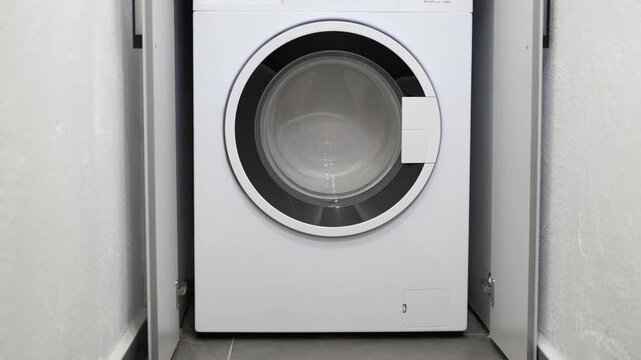 White washing machine spinning clothes in a laundry room. Modern white washing machine with a black circular door starting its washing cycle, with the drum full of clean laundry spinning inside