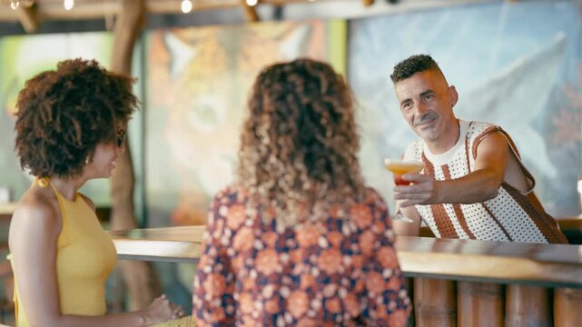 Friendly bartender serving a delicious colorful cocktail to two female friends sitting at a wooden counter in a vibrant tropical themed bar