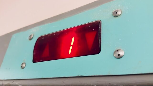 Red LED elevator floor indicator showing blinking digit and arrow on turquoise metal panel, screw heads visible, glass window