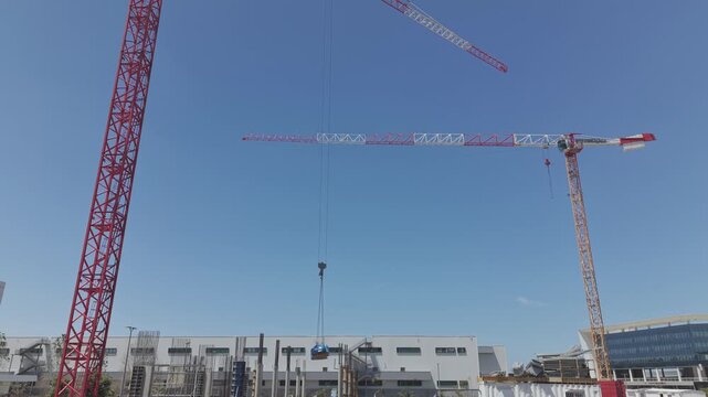 Two tower cranes lowering heavy blue load at Israeli urban construction site against clear blue sky
