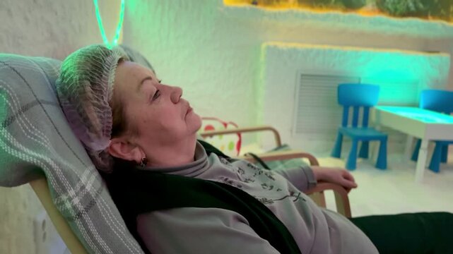 Elderly woman reclining in salt cave halotherapy chamber wearing cap on recliner under halogen neon lighting salt encrusted walls