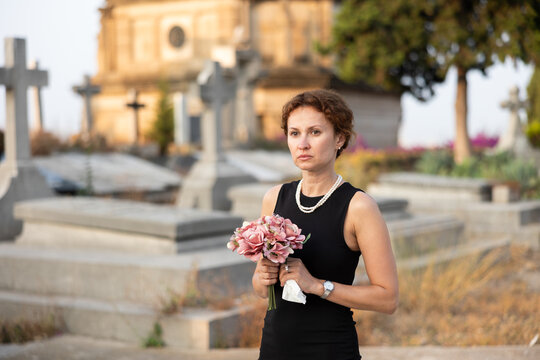 Sorrowful young woman in black dress with bouquet of flowers and handkerchief in hands, stands in cemetery. Visiting grave of deceased relative, mourning dead loved one. Appeal to God, prayer for dead