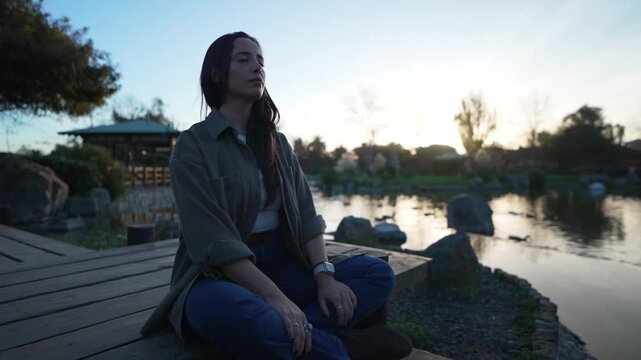 Young woman meditating in a serene park, sitting by a tranquil lake during a beautiful sunset