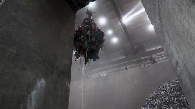 A waste handling claw suspended above a large accumulation of mixed material inside a waste processing plant. The mechanical arm holds a compact mass of discarded items drawn from the pit below. 