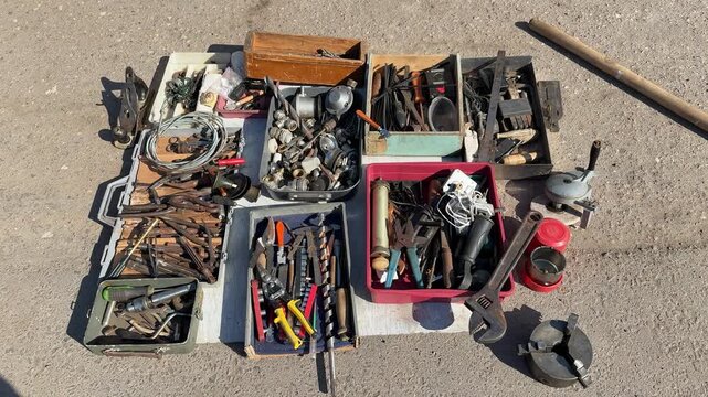 Tools for sale on asphalt showing assorted pliers, wrenches, screwdrivers and sockets in open boxes jars of screws and rusty