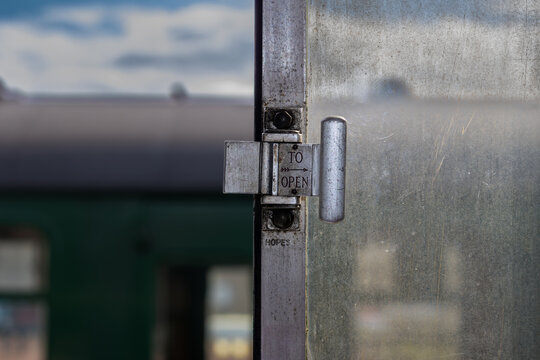 Close-up of a metal window latch with &ldquo;to open&rdquo; instruction on an old railway carriage, showing worn glass, metal texture, and blurred train exterior in the background.