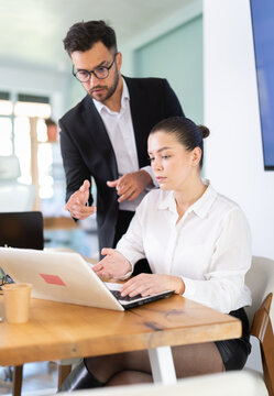 IT specialist man helps a young female employee work on a laptop against the backdrop of the interior of a modern office. Team of web designers is working on a new website