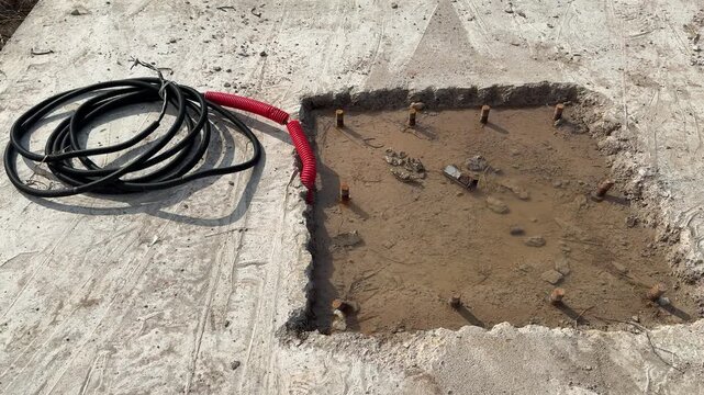 Muddy foundation pit with anchor bolts, water pooling in sandy excavation, red safety rope along perimeter, exposed bolt studs