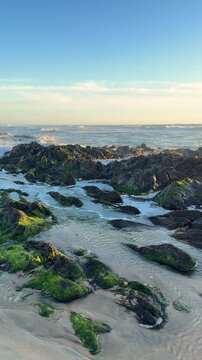 SLOW MOTION SHOT - Sea, beach boulders, pebble shore, and waves at sunset. Sea waves breaking on rocks. Sunny beach with dunes and blue sky in Esposende, Portugal. 