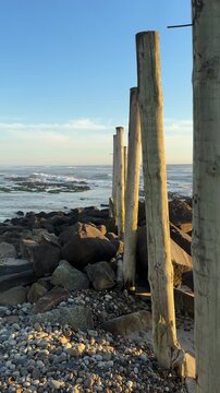 SLOW MOTION SHOT - Wooden stakes on the beach in Esposende, Portugal. Sea, beach boulders, pebble shore, and waves at sunset.