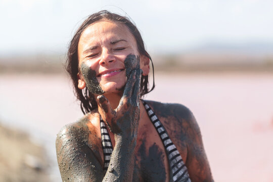 Woman smiling and applying black mud to face as part of natural skincare ritual by pink lake