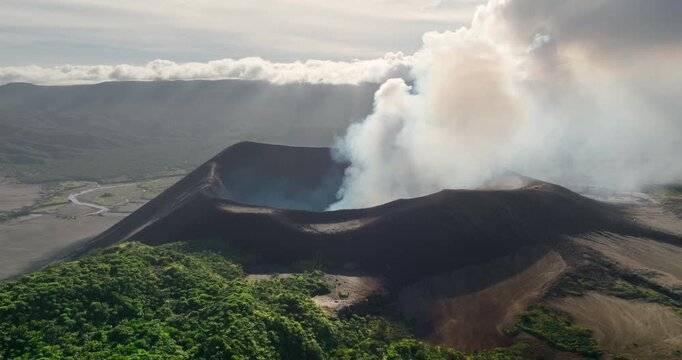 Mount Yasur on Tanna Island, Vanuatu, actively venting steam volcano and ash from its crater, surrounded by volcanic soils and green vegetation beneath a cloudy sky. Aerial view drone flight panorama