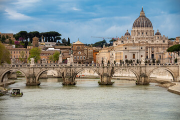 Fototapeta premium View on stone-arched bridge on Tiber river and St Peter's Basilica in Rome, terracotta buildings, domes and umbrella pines rise on gently sloping Vatican hill, classic Roman skyline