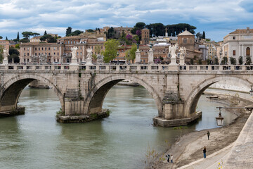 Fototapeta premium View on stone-arched bridge on Tiber river in Rome, balustrade, marble statues, terracotta buildings, domes and umbrella pines rise on gently sloping Vatican hill, classic Roman skyline