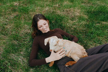 Fototapeta premium Happy woman lying on grass beside a playful goat kid, embracing nature and friendship in an outdoor lifestyle moment, warm sunlight, relaxed mood, gentle bonding between human and animal