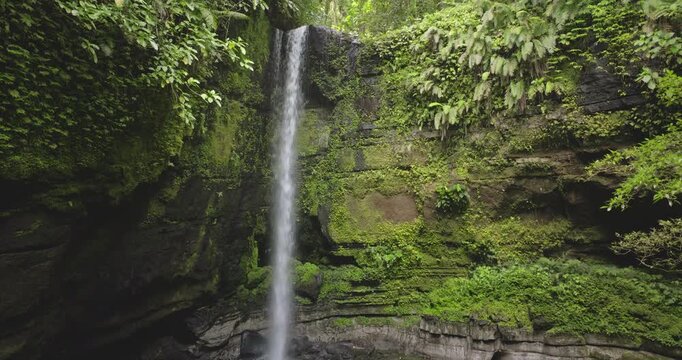 Drone footage capturing a tropical waterfall cascading over mossy rock walls into a clear pool surrounded by lush rainforest vegetation and vibrant green foliage, pristine nature of Solomon islands