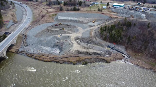 Cinematic drone view of infrastructure construction along the Salmon River Mitis with brown floodwaters, heavy machinery and traffic crossing the existing bridge in Quebec, Canada, 2026.