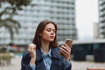 Fototapeta premium Young woman with long brunette hair wearing a dark coat looks thoughtfully at her smartphone while standing in an urban environment with blurred buildings in the background.