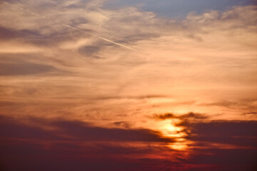 Obraz premium Golden sunrise through cloudy sky with sunbeams and a jet contrail crossing from top left to right.